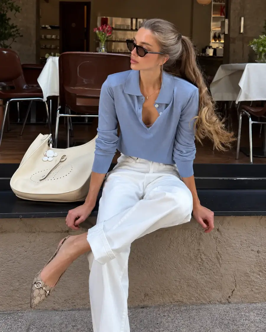 Woman wearing a polished light blue knit top with white tailored trousers, sunglasses, pointed flats, and a cream handbag, illustrating a simple, well-fitted outfit that creates a wealthy, elegant look.
